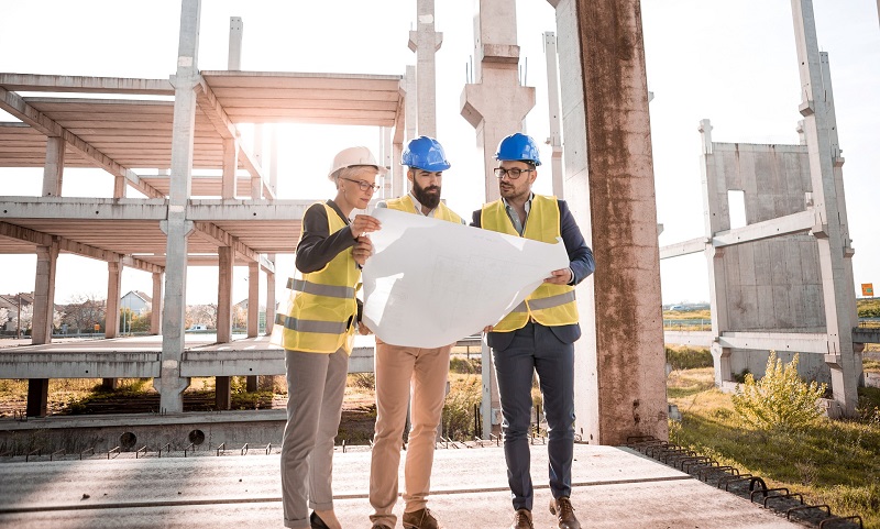 Applying Lean At Massive Scale Three People in Construction Safety Gear Looking at Blueprints